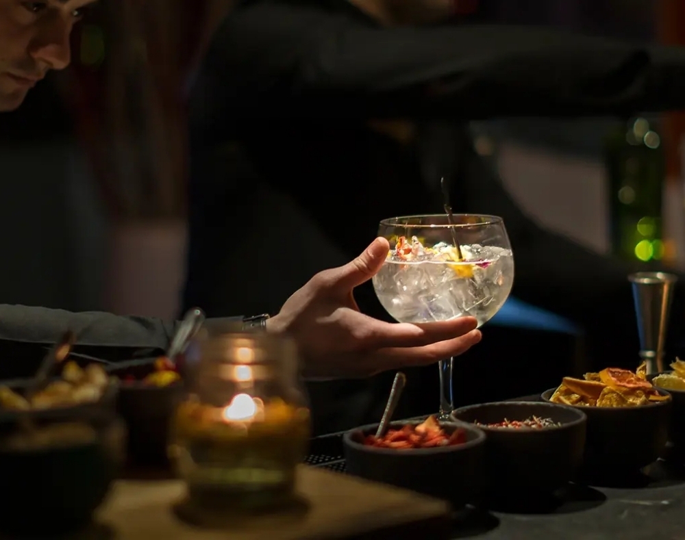 Bartender preparing a cocktail in a large goblet with ice and edible flowers, dim candlelit bar with bowls of garnishes.