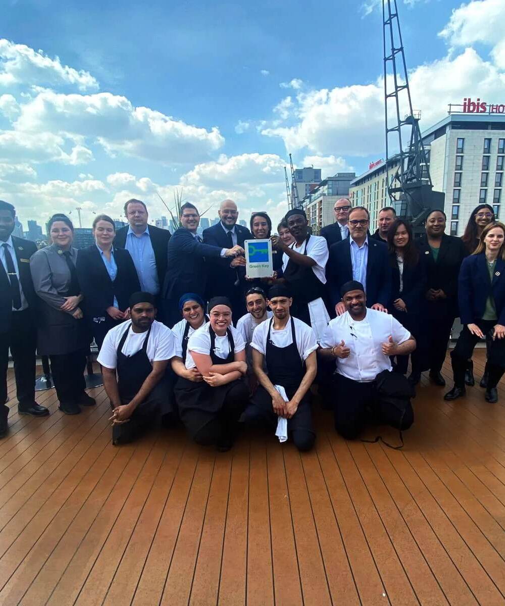 Hotel team of chefs and staff on a waterfront rooftop terrace holding a Green Key award, ibis hotel and skyline behind.