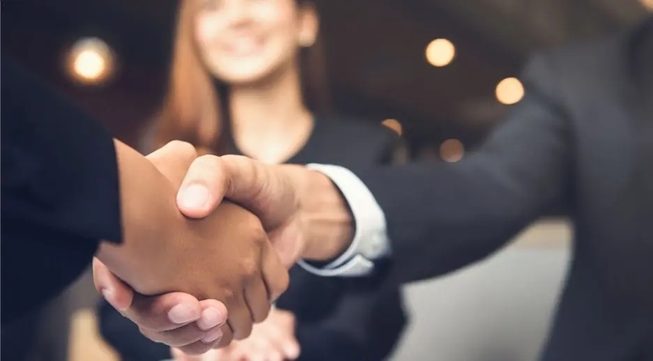 Hotel staff and guest shaking hands at reception in modern lobby, smiling concierge in background, warm professional welcome.