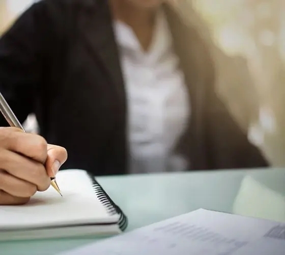 Hotel business-center staff writing in a spiral notebook at a glass desk, paperwork nearby, dressed in professional attire