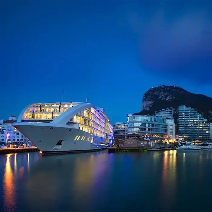 Luxury multi-deck yacht docked by waterfront hotels and glass buildings, lit balconies and mountain backdrop at dusk reflected in water.
