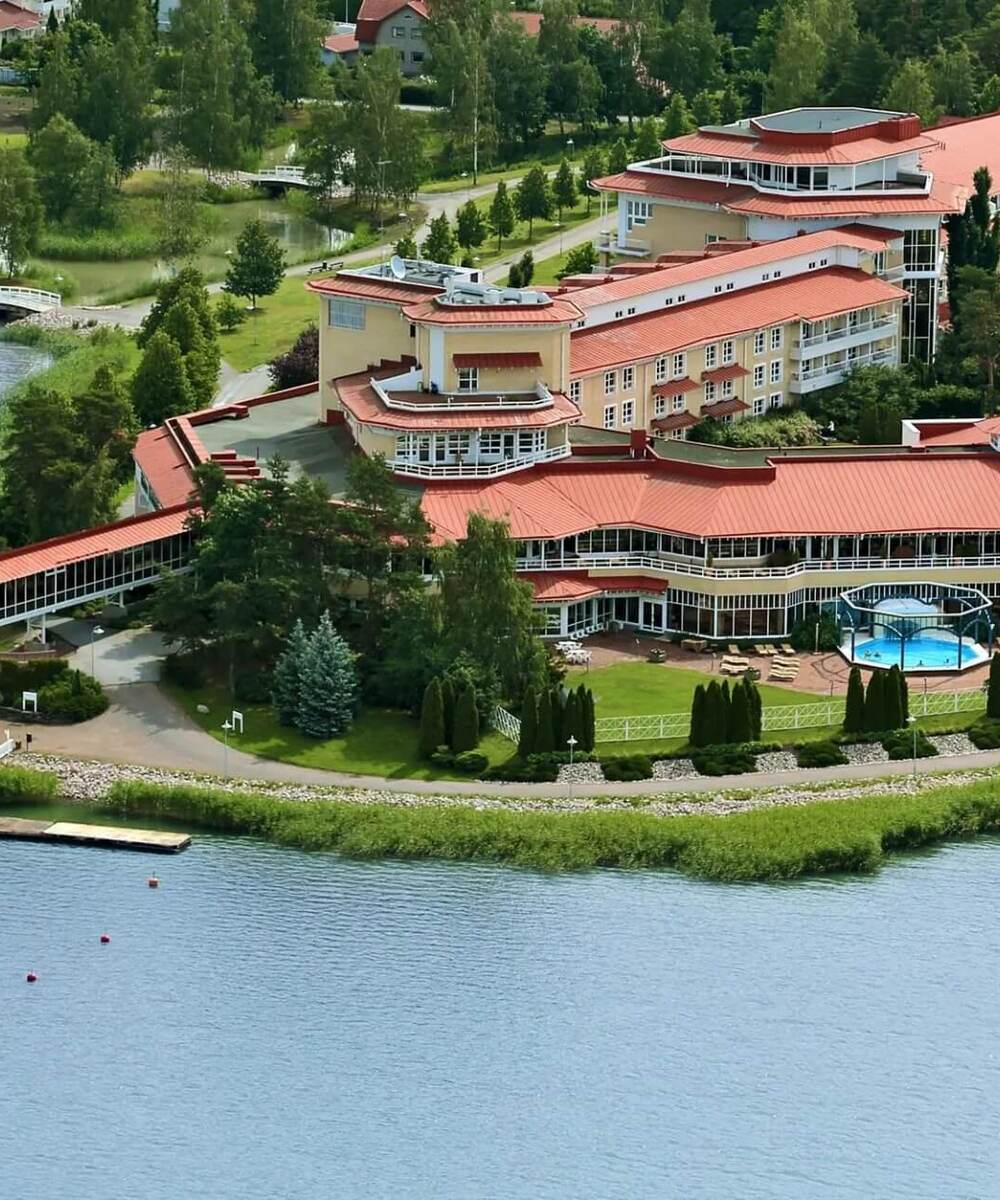 Lakeside resort with red‑roofed hotel, waterfront pier, outdoor pool and sun terraces framed by lawns, trees and walking paths