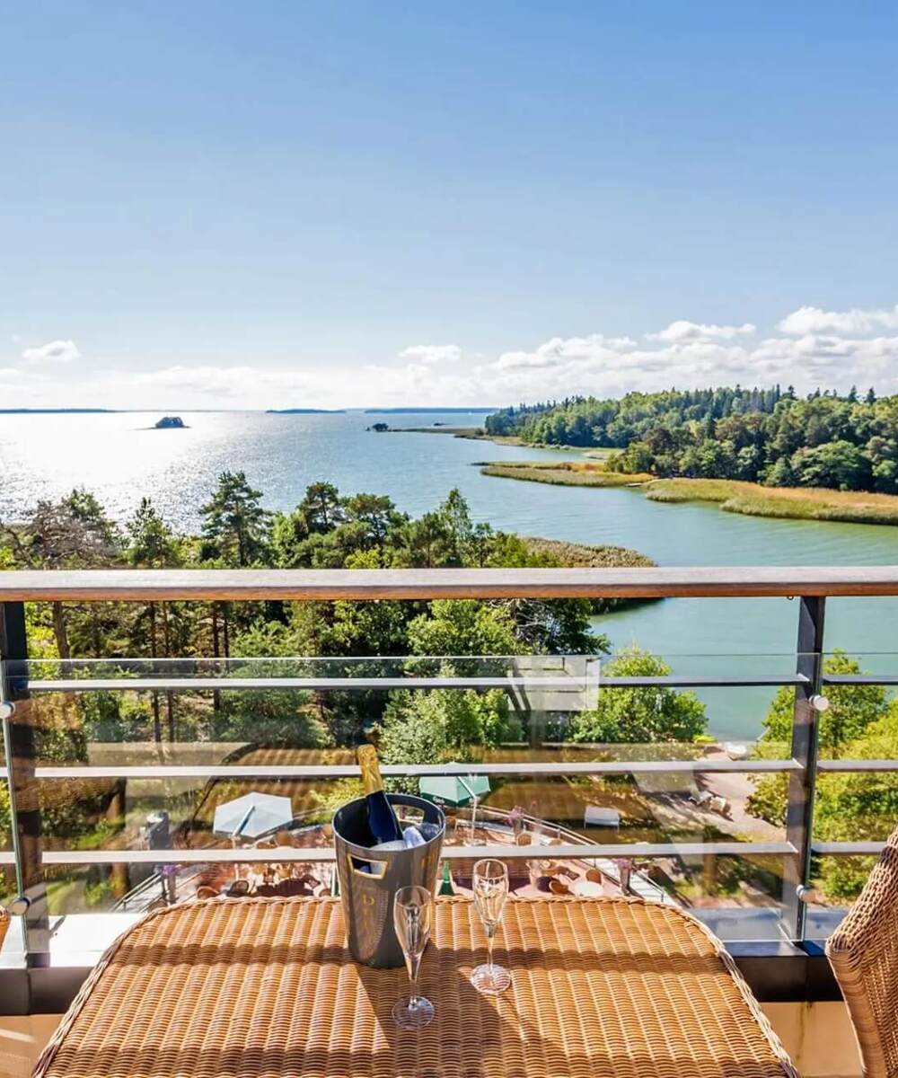 Private balcony with wicker chairs and chilled champagne on a table, overlooking sunlit archipelago, green shoreline and sea.