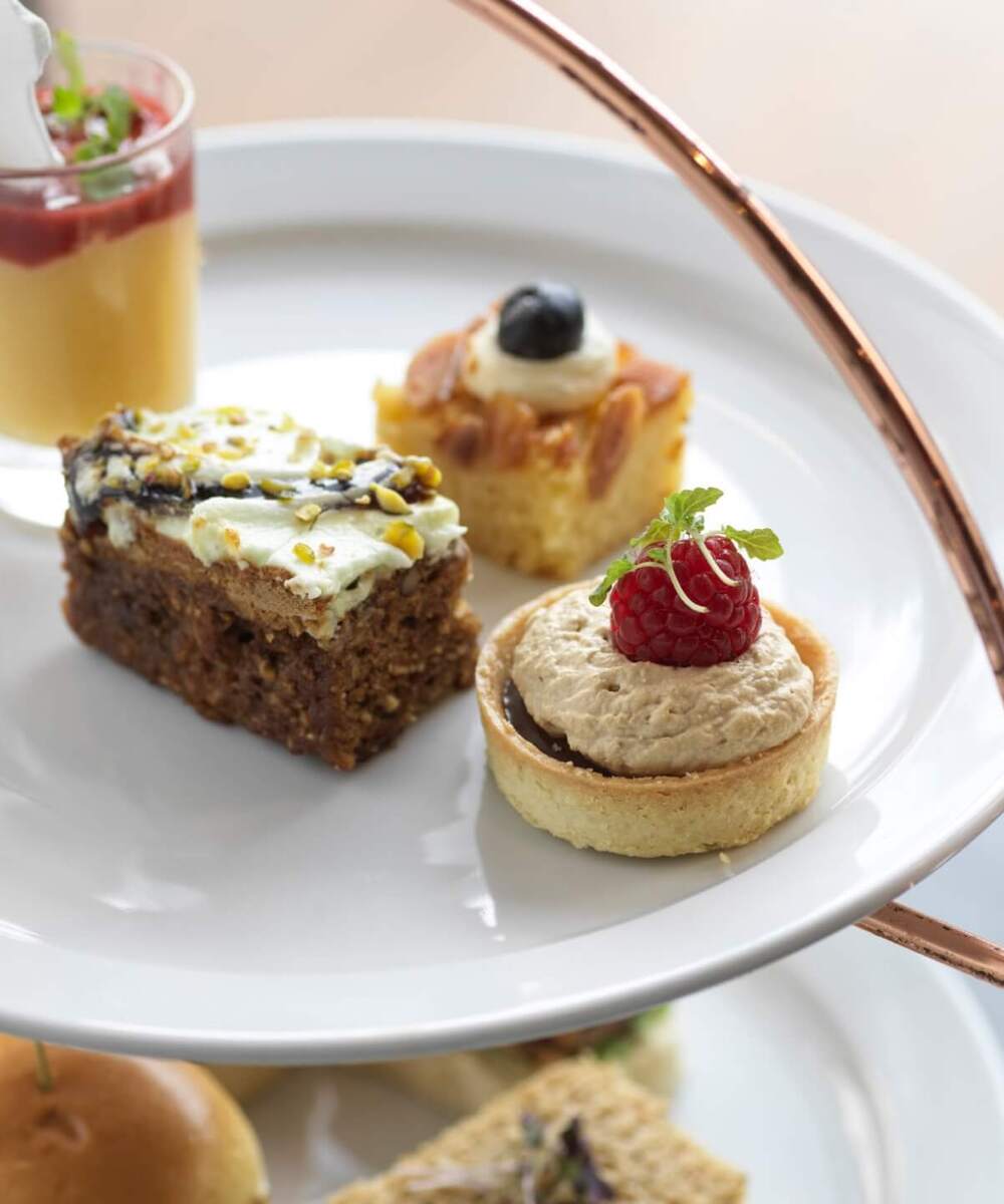Afternoon tea pastries on a white tiered stand: raspberry tart, pistachio cake, almond square and custard shot