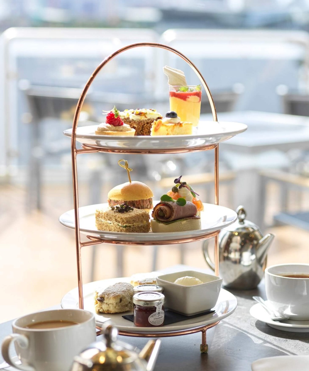 Three-tiered afternoon tea stand with scones, finger sandwiches, pastries and dessert glass beside teapot on sunlit terrace.