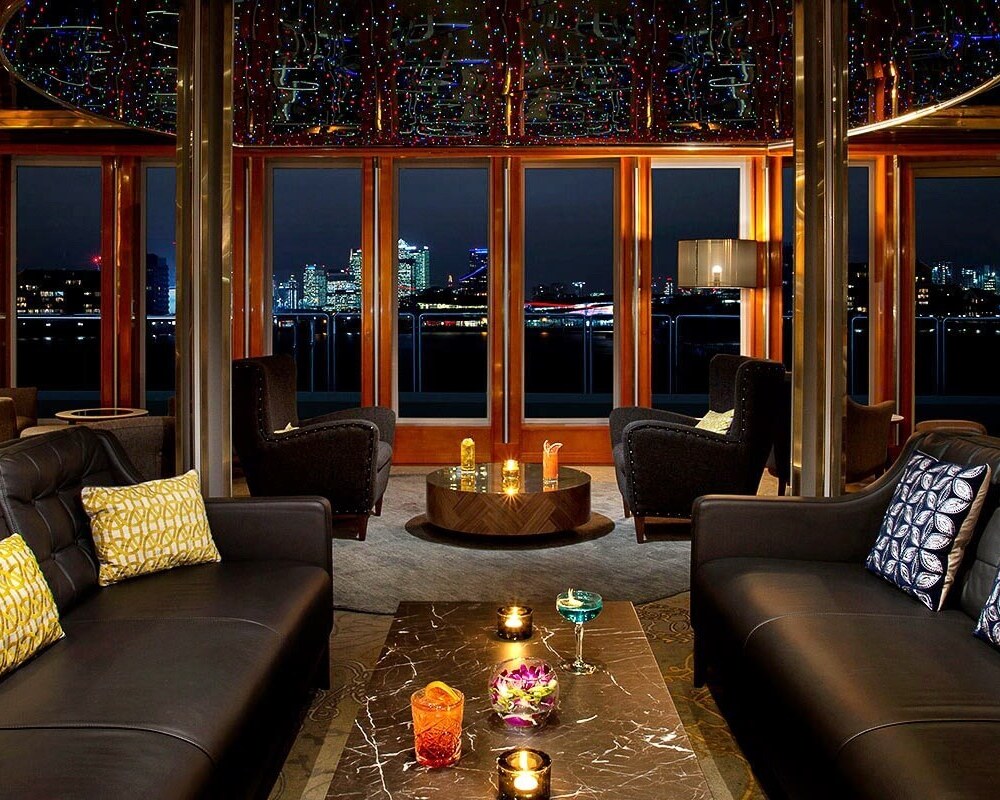 Hotel lounge at night with black leather sofas, patterned cushions, cocktails on a marble table and city skyline views.