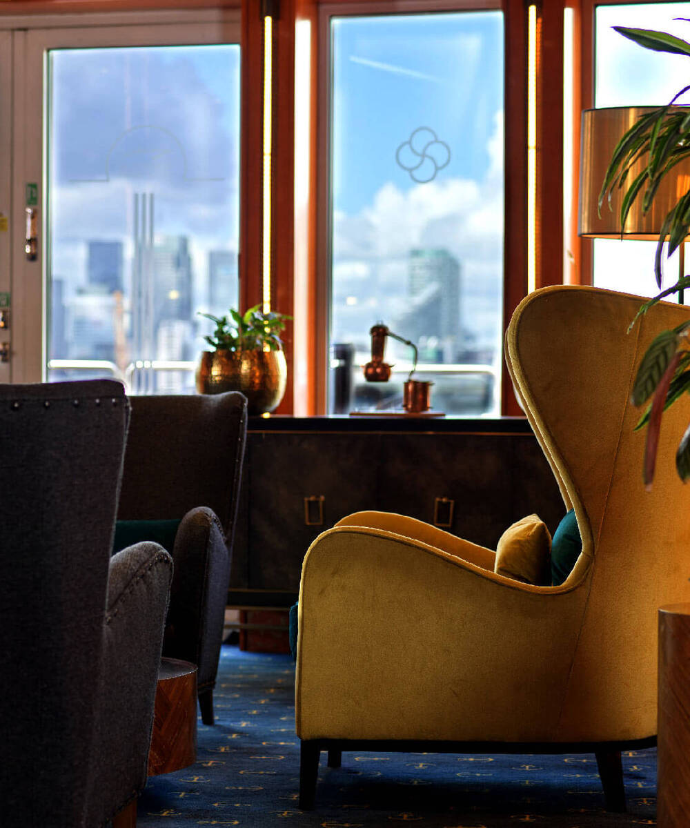Cozy hotel lounge with velvet armchairs, plants and warm lamp by large windows showing a blurred city skyline