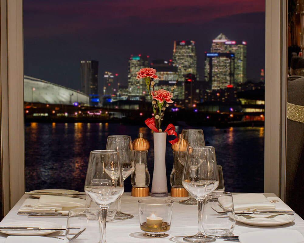 Candlelit waterfront table with white linen, crystal glassware, pink flowers and gold-banded high-back chairs, city skyline at night