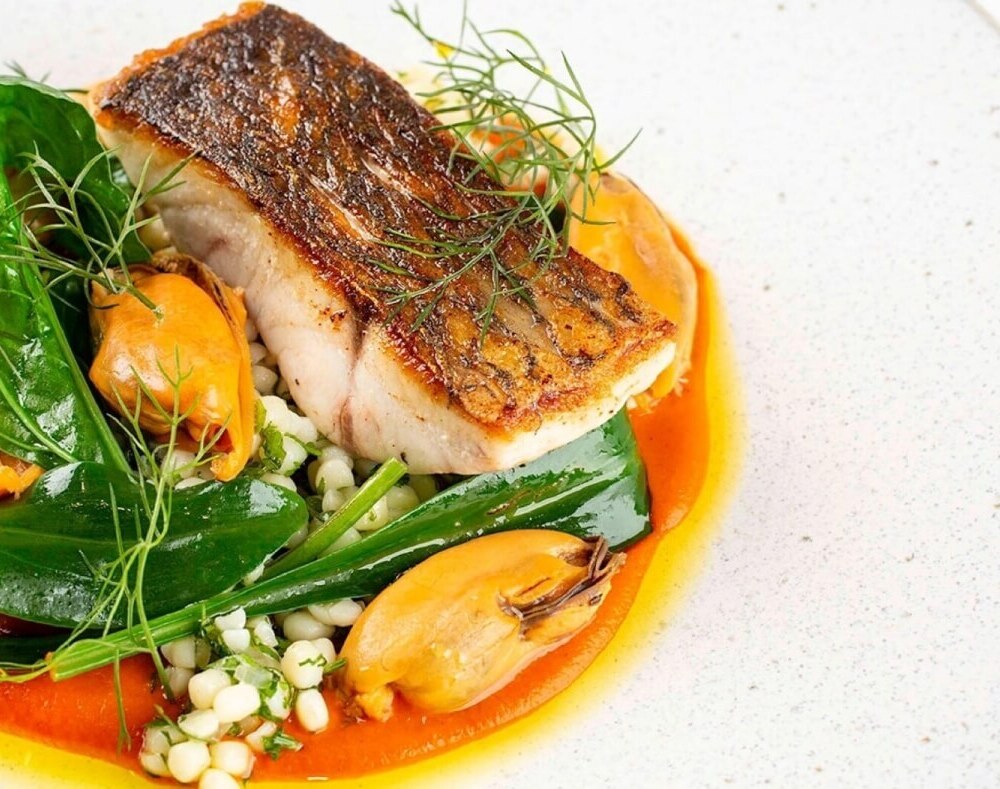 Seared fish fillet with mussels, pearl couscous, wilted greens and red pepper sauce, plated for hotel fine-dining.