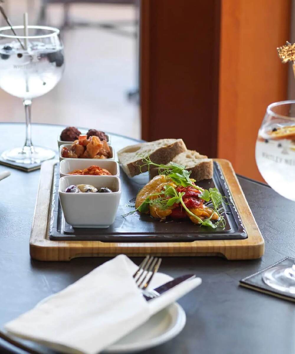 Tapas sharing board with olives, dips, roasted peppers and bread on a wooden tray, served with two gin-and-tonic cocktails.