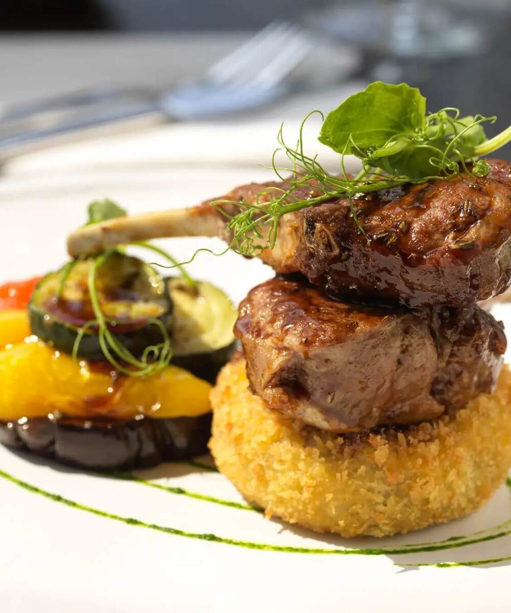 Two glazed lamb chops stacked on a golden breadcrumb croquette, served with grilled vegetables and pea-tendril garnish.