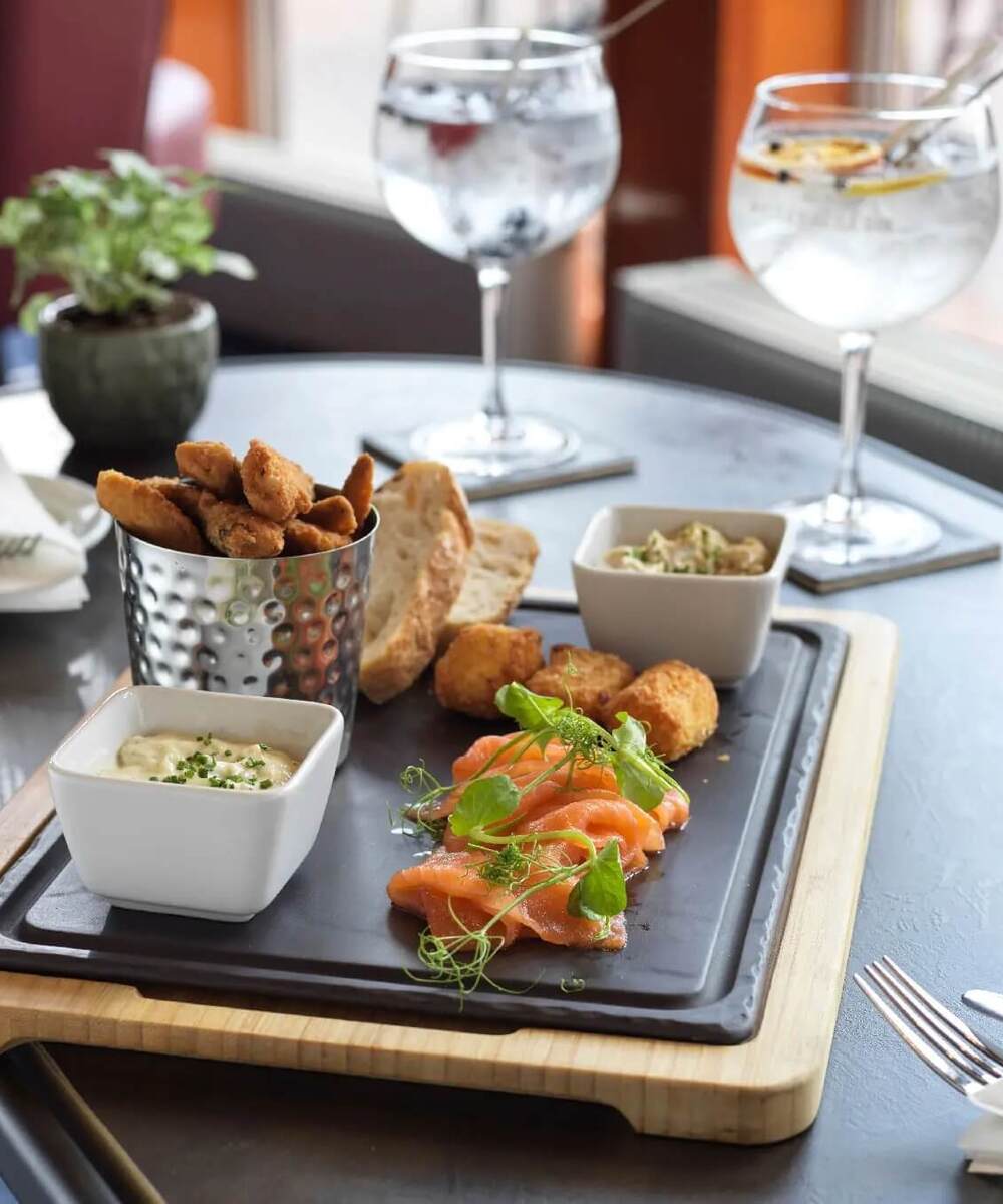 Smoked salmon and fried bites on slate board with dips, crusty bread and two gin-and-tonic glasses at hotel bar.