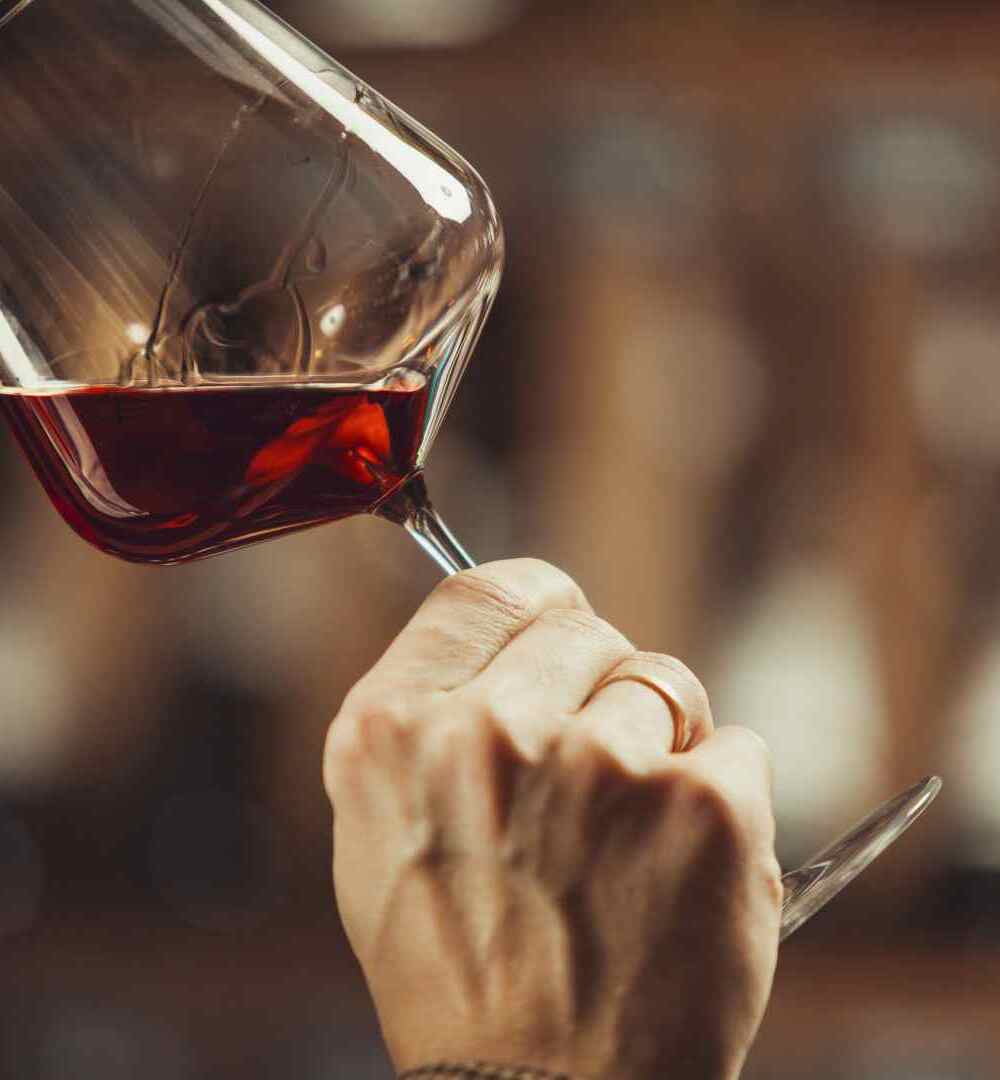 Guest smelling red wine at hotel wine bar, close-up of glass held to nose, hand with ring, warm wood-paneled bokeh lighting