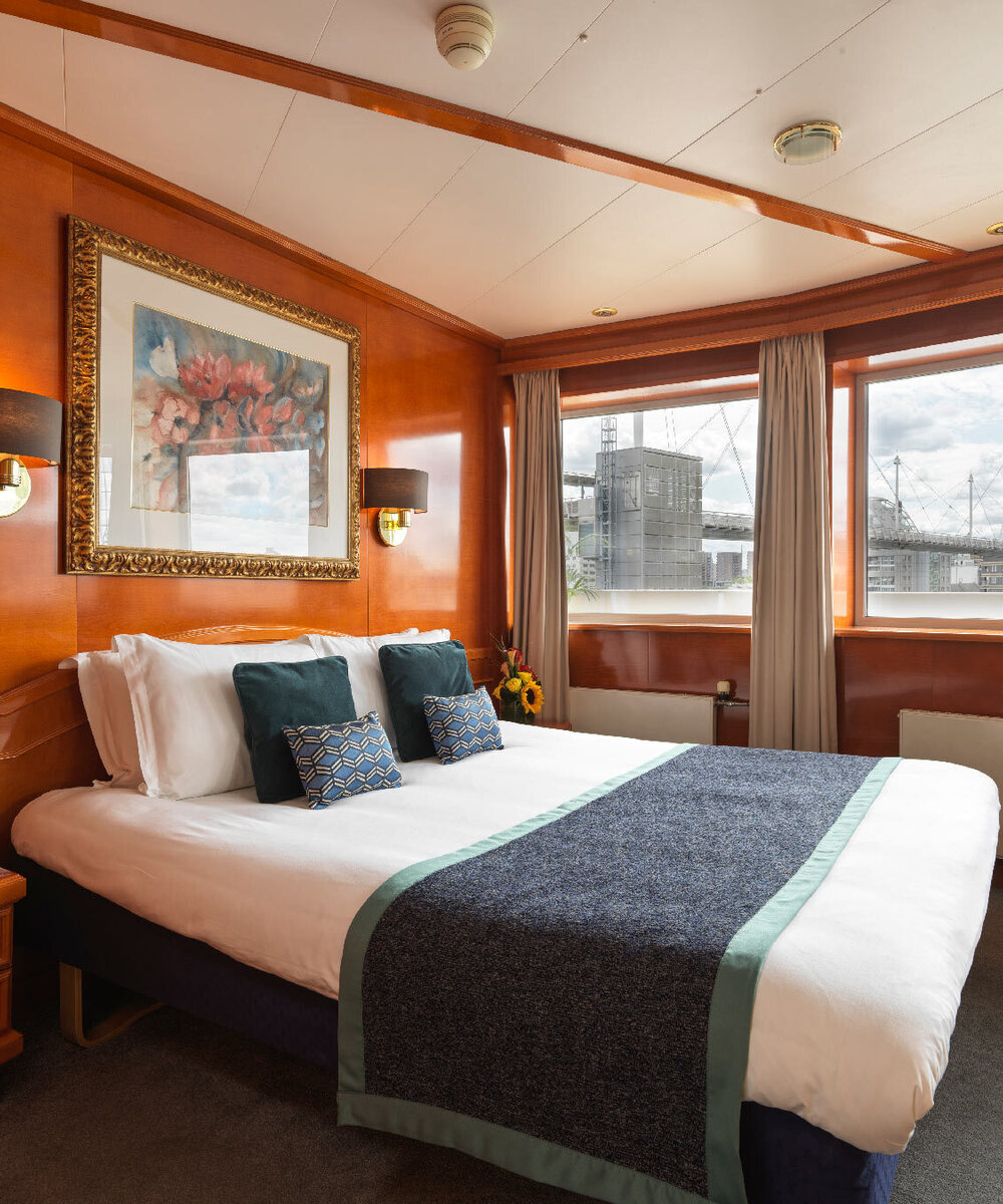 Wood-paneled double room with king bed, patterned cushions, framed art and bedside lamps; windows with modern bridge and city view.