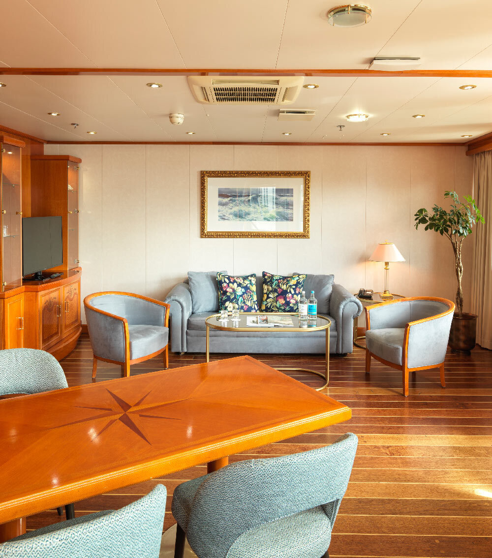 Luxury suite lounge with warm wood finishes, dining table, plush sofa and chairs, coffee table, lamp and sunlit windows.