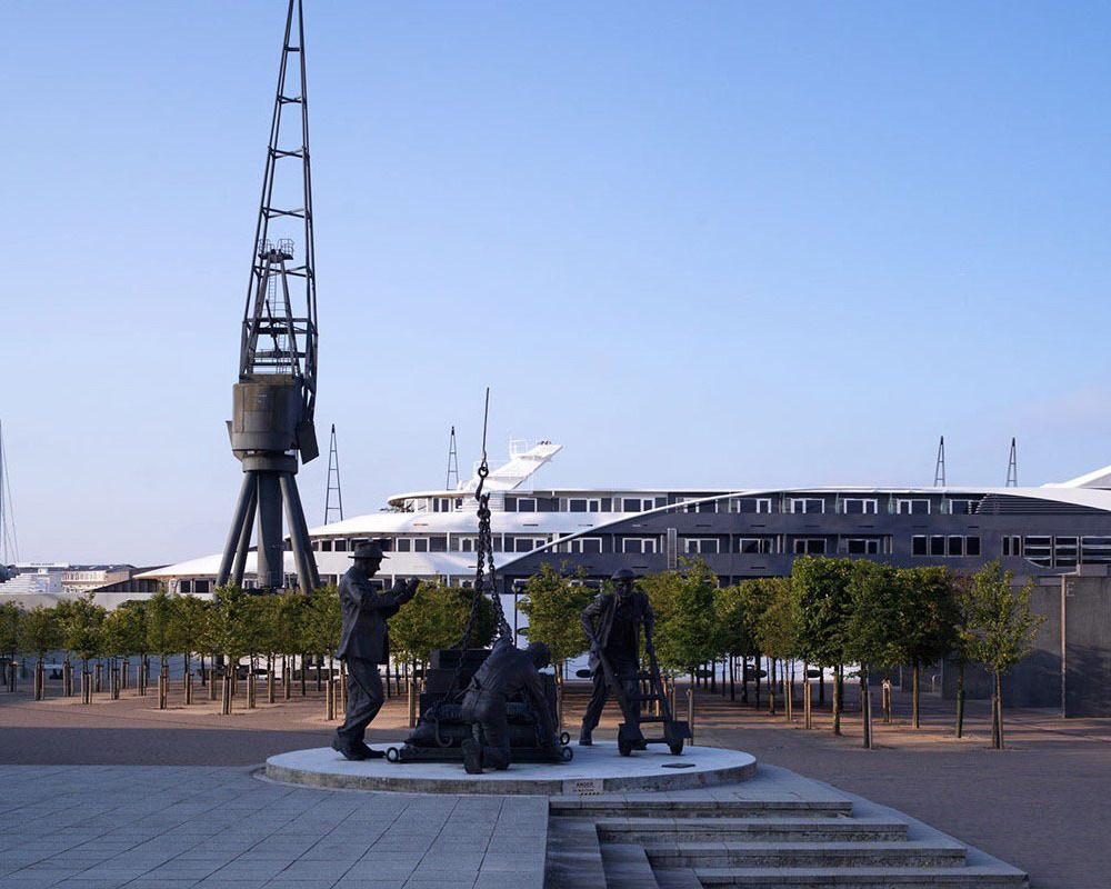 Waterfront promenade with tree-lined walkway, bronze dockworker statue, moored white superyacht and tall industrial cranes.