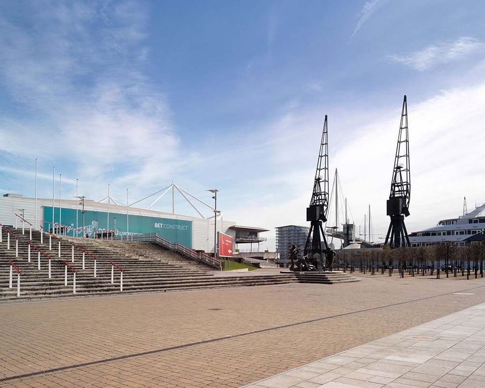 Waterfront ExCeL London convention center with broad stone steps and glass atrium, dockside cranes and moored yachts on a sunny plaza