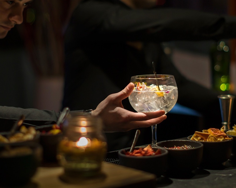 Bartender in black serving a large iced cocktail with citrus garnish at a candlelit hotel bar with bowls of snacks.