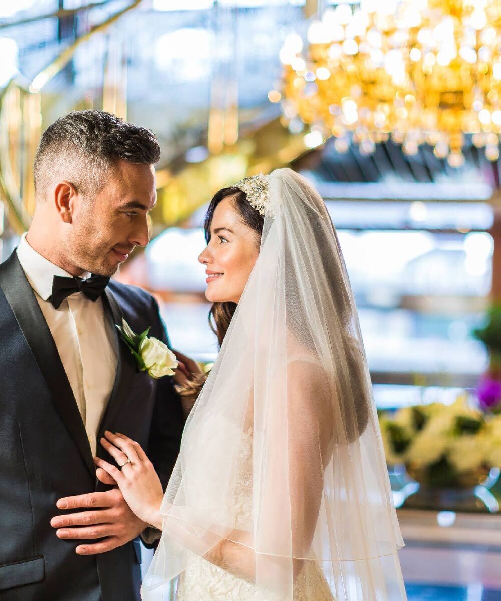 Bride and groom smile in hotel lobby beneath a crystal chandelier, surrounded by wedding flowers and a sweeping staircase
