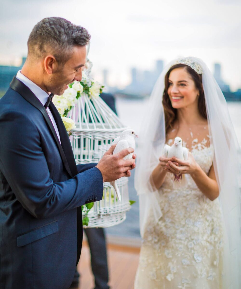 Bride in lace gown and groom in tuxedo release white doves on a waterfront terrace with a city skyline and floral birdcage.