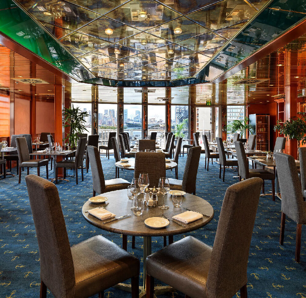 Elegant hotel dining room with mirrored geometric ceiling, polished wood panels, set tables, upholstered chairs and waterfront city skyline view.