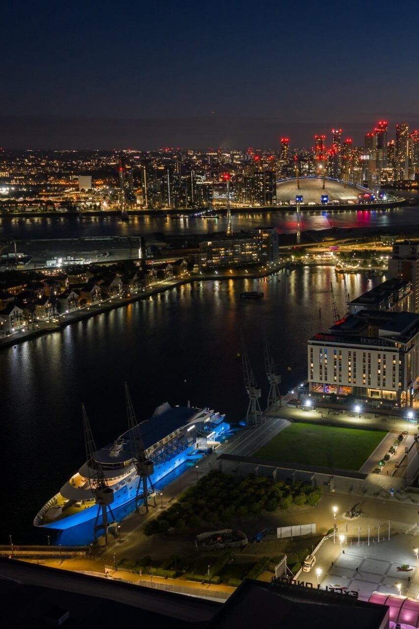 Riverside hotel and marina lit at night with illuminated arena dome and glittering city skyline across the water.
