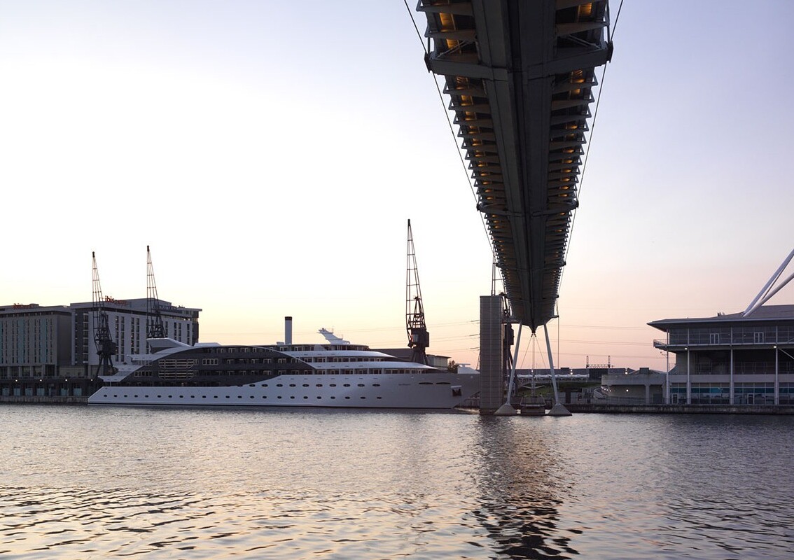 Luxury yacht moored beside modern waterfront hotels and a riverside conference center under an elevated pedestrian bridge at dusk.