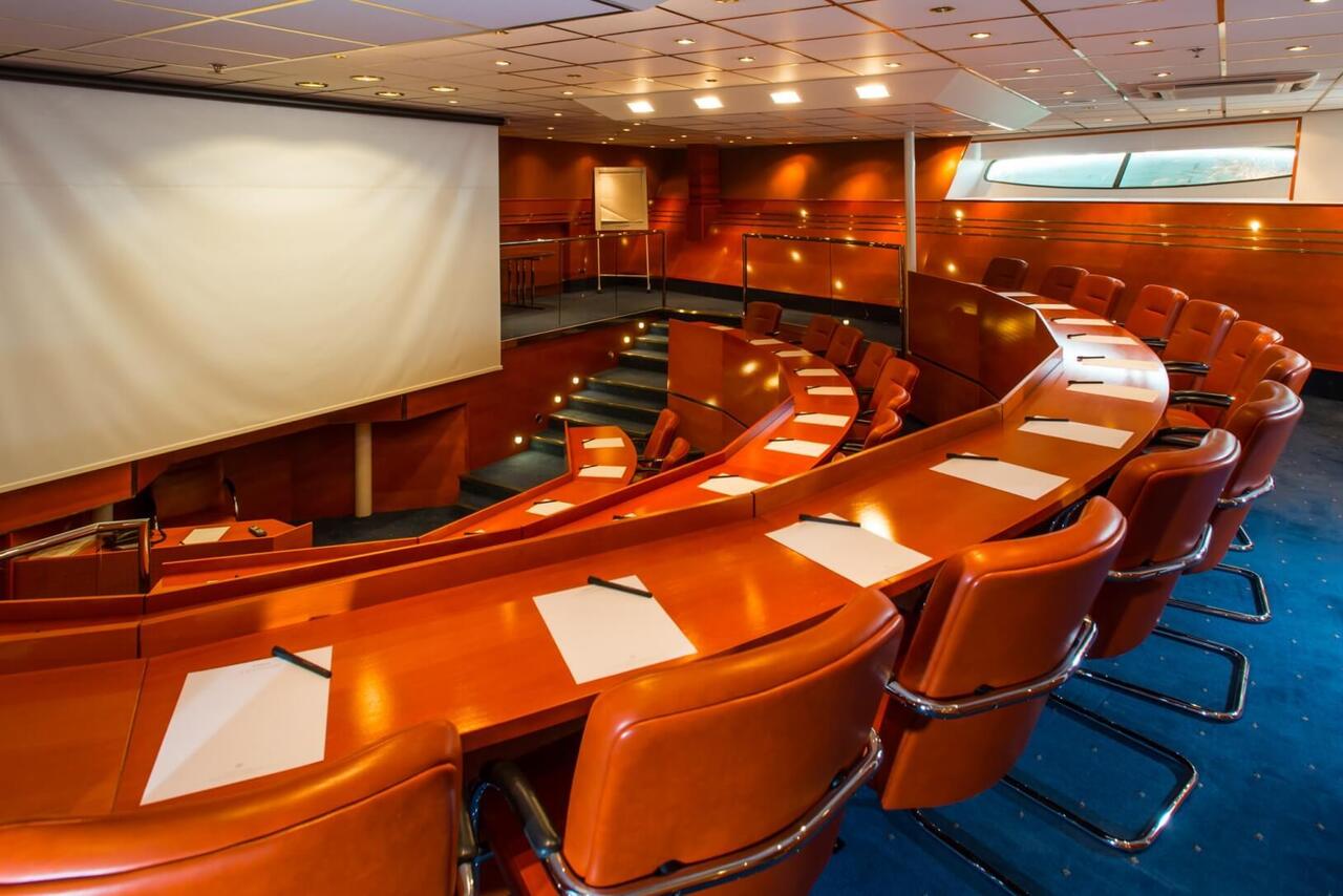 Tiered conference theater with large projection screen, curved wood desks, leather swivel chairs, pens and pads, soft lighting and narrow windows.