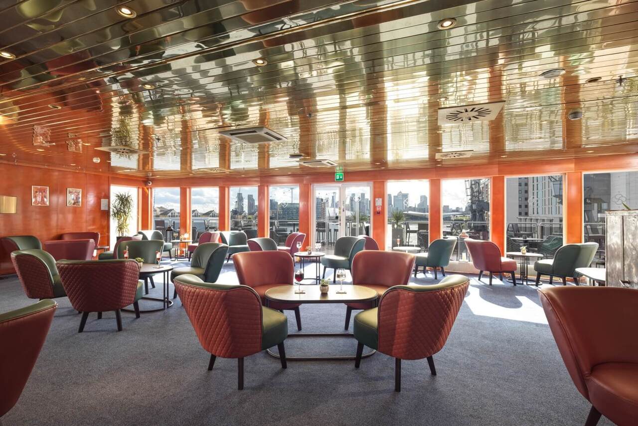 Sunlit hotel lounge with quilted armchairs, tables with drinks, reflective ceiling and city skyline terrace view