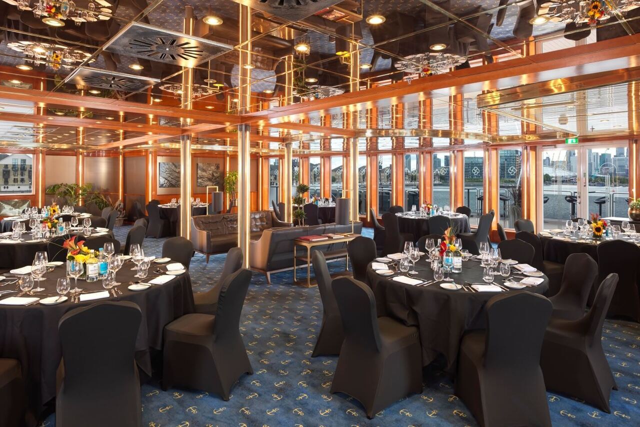 Elegant waterfront banquet room with round black-clothed tables, formal place settings, mirrored ceiling and city skyline views.