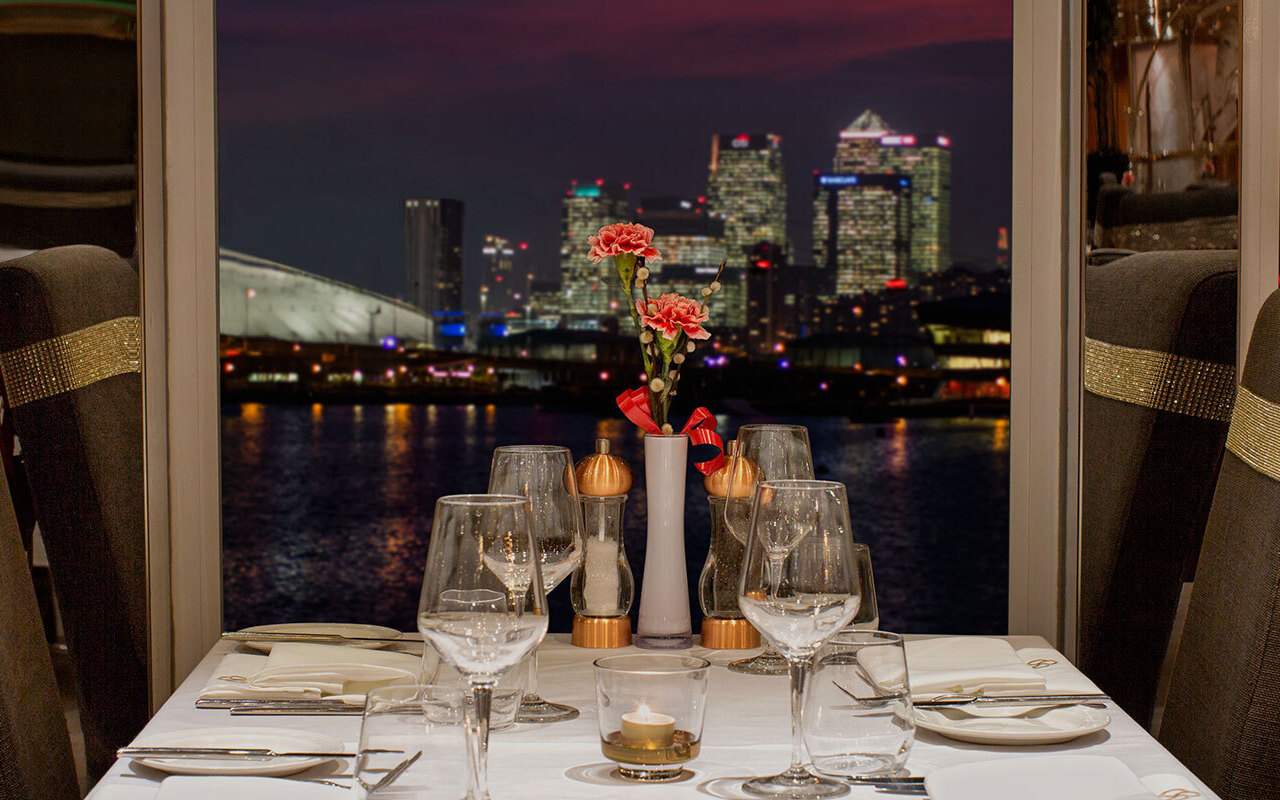 Candlelit waterfront table with white linen, crystal glassware, pink flowers and gold-banded high-back chairs, city skyline at night