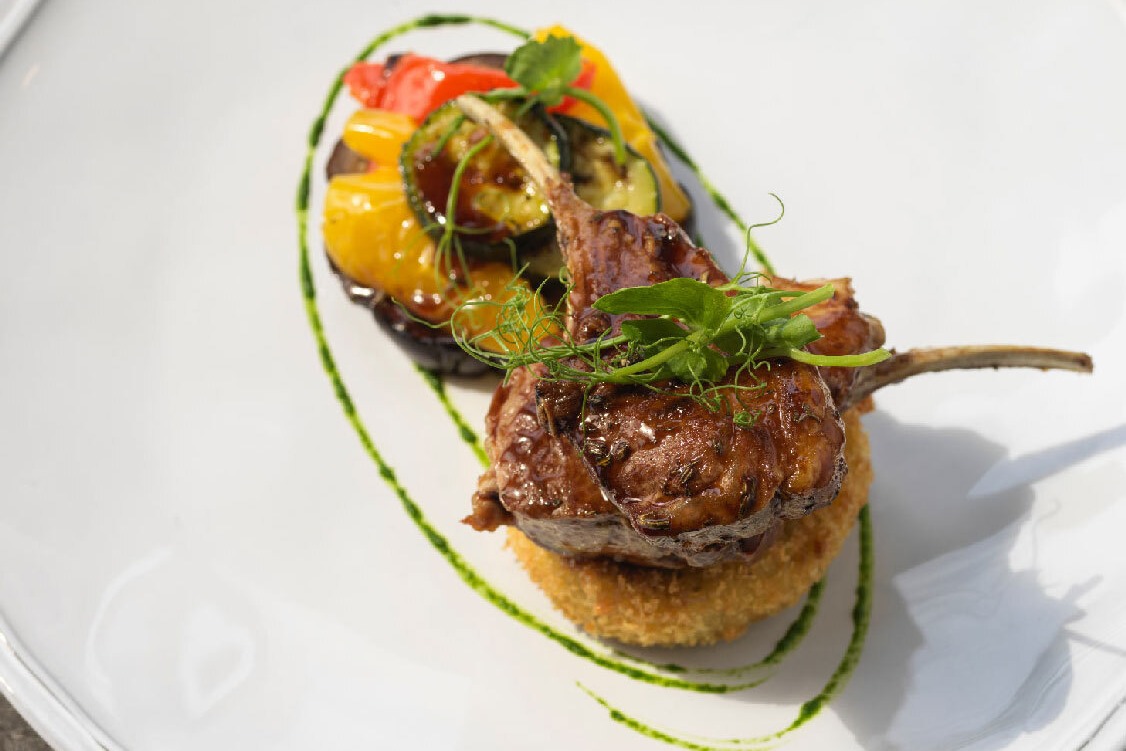 Seared lamb chops on a crispy potato cake with roasted cherry tomatoes and zucchini, herb oil swirl on white plate