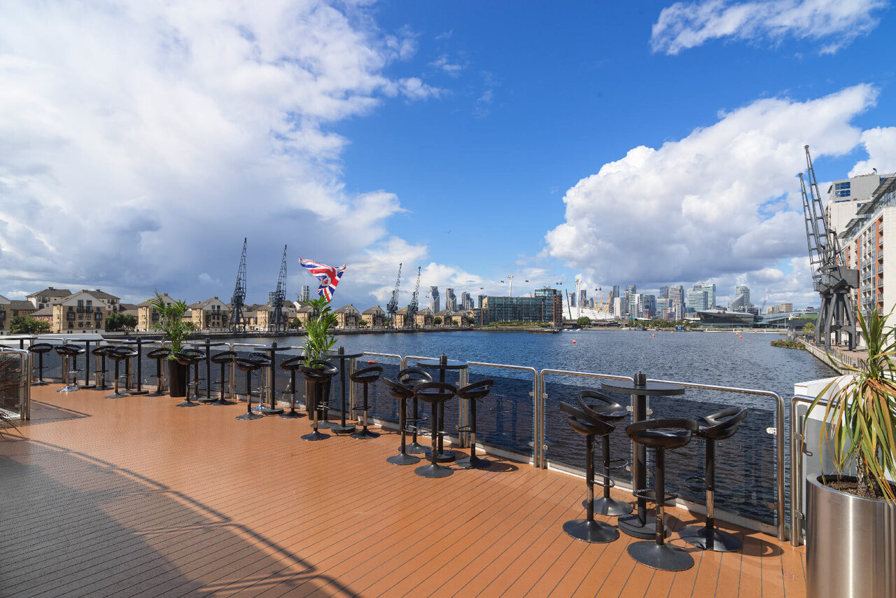 Riverside hotel terrace with bar stools, plants and panoramic dock view of historic cranes, city skyline and a Union Jack flag