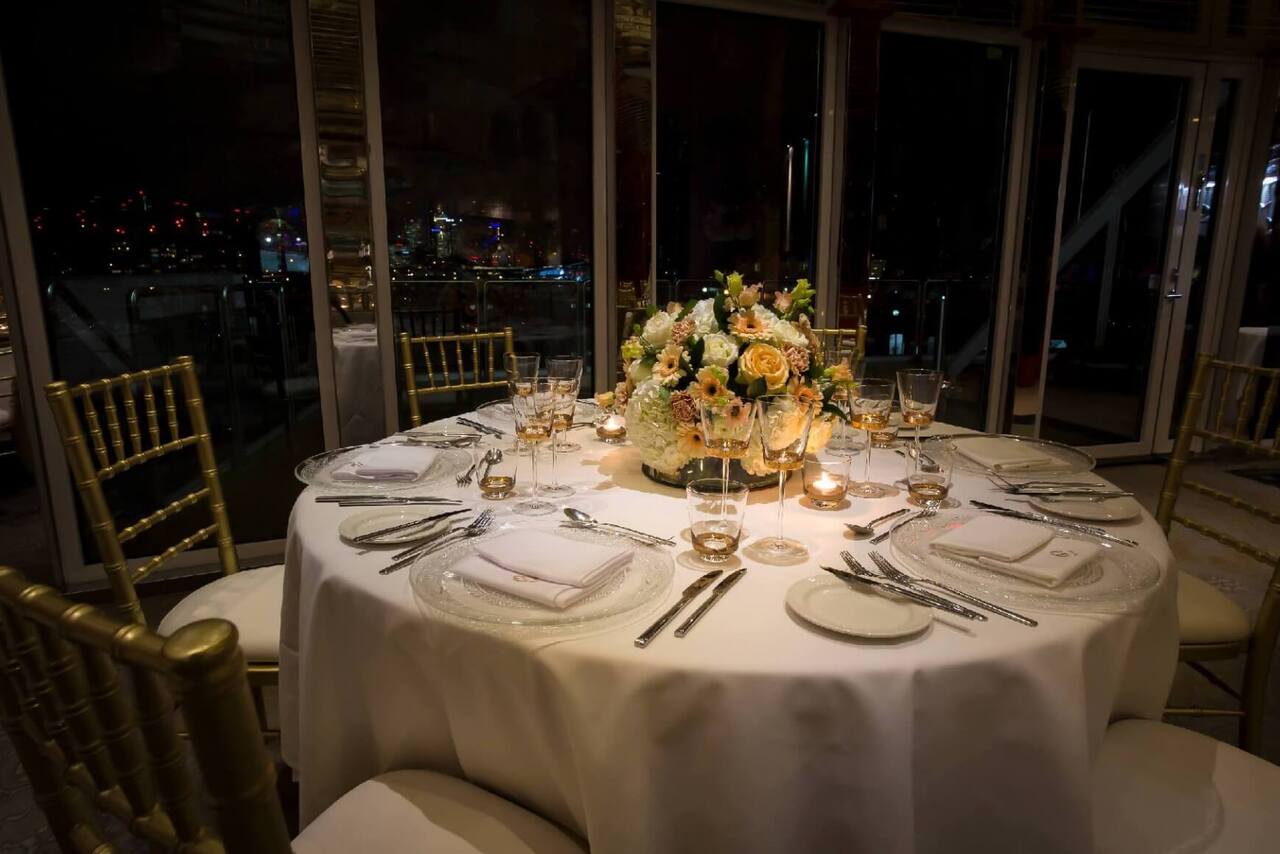 Round table set for six with white linen, crystal plates, gold glassware and pale floral centerpiece, gold chairs, city night view.
