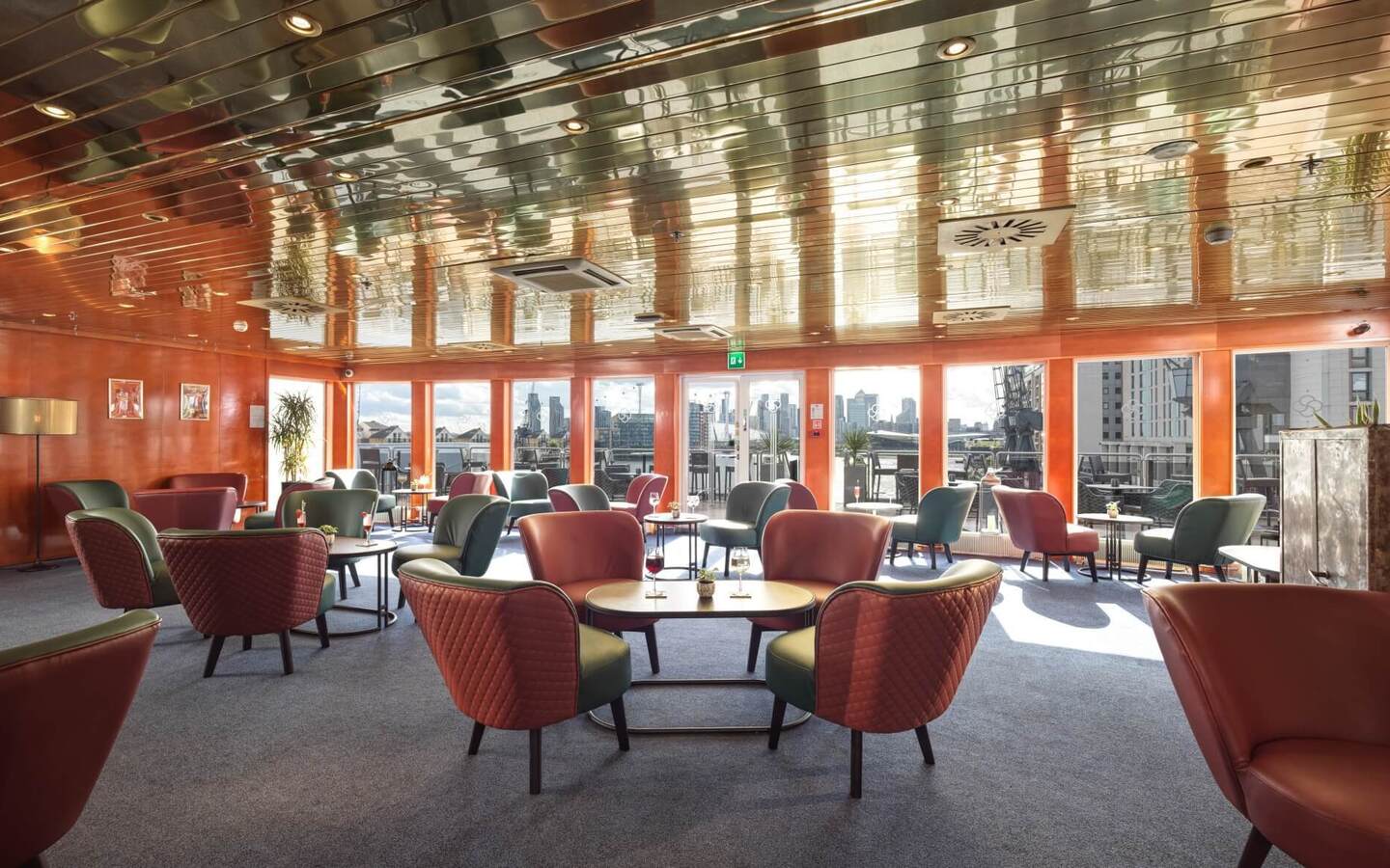 Sunlit hotel lounge with quilted armchairs, tables with drinks, reflective ceiling and city skyline terrace view