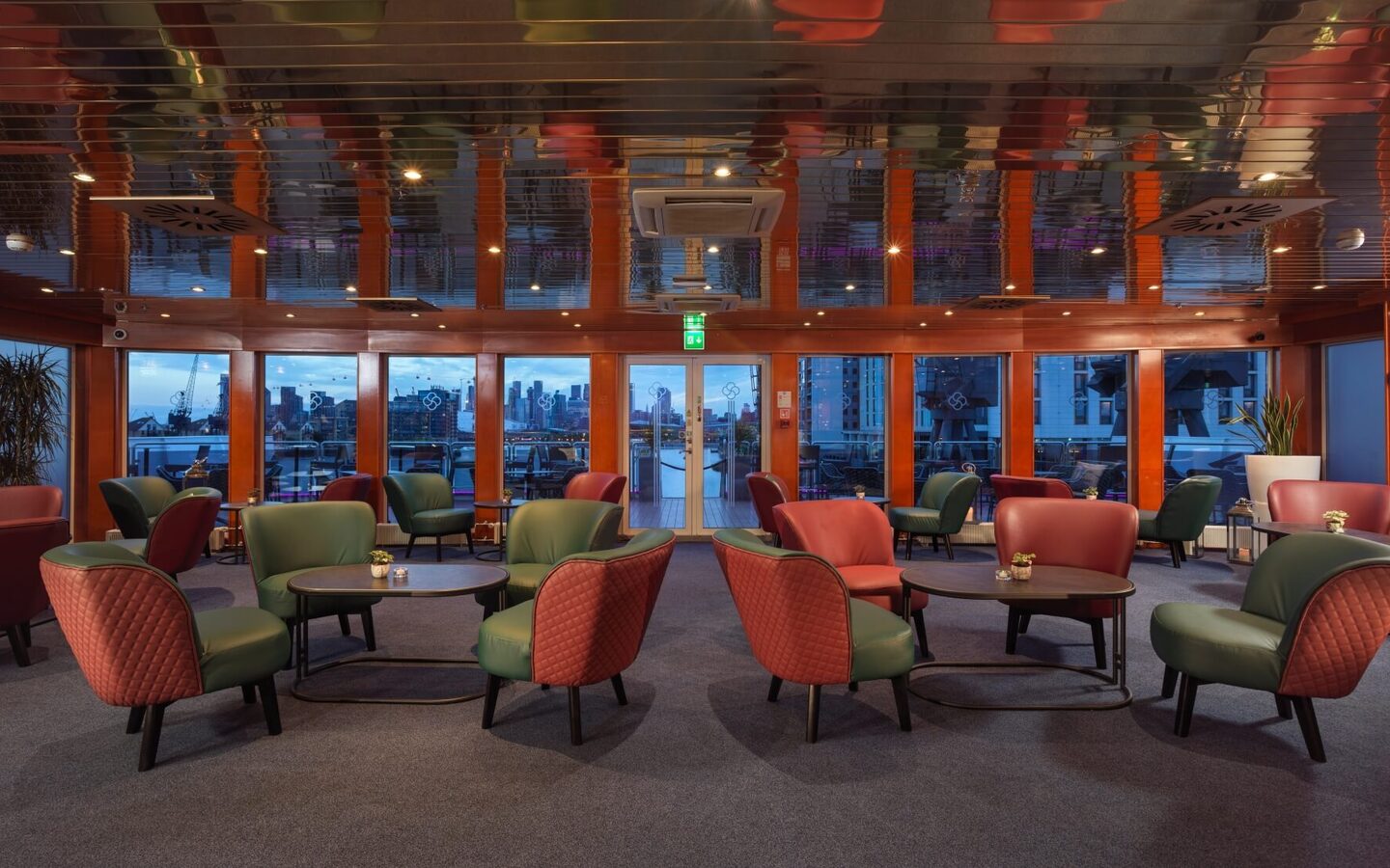 Hotel lounge with green and red quilted armchairs, low tables, reflective ceiling and glass doors to a waterfront terrace and city skyline.
