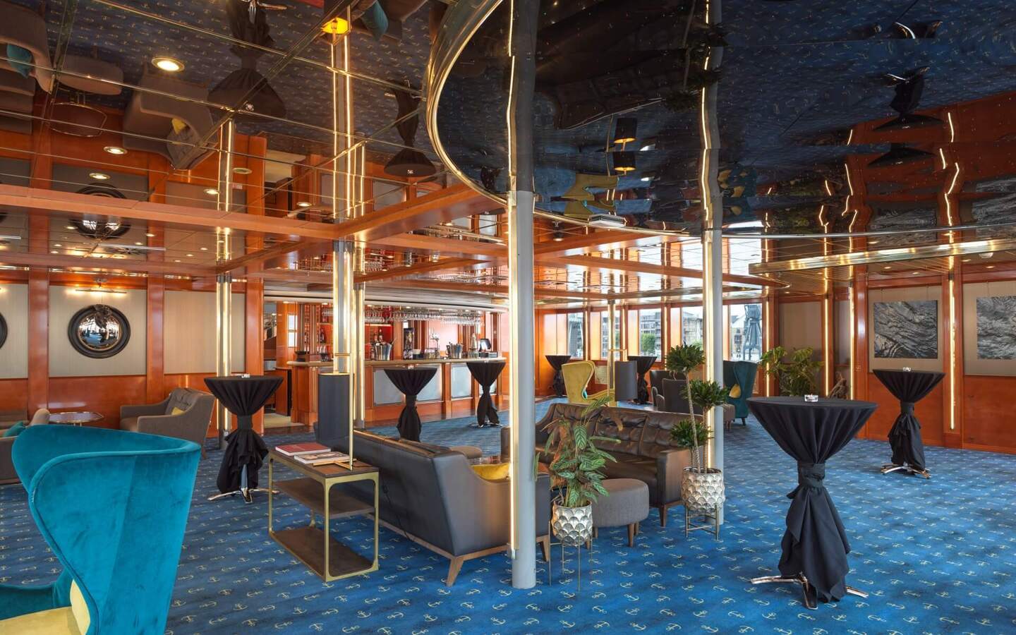 Nautical-themed lounge with mirrored ceiling, warm wood beams, bar, teal chairs and sofas, anchor-patterned blue carpet.