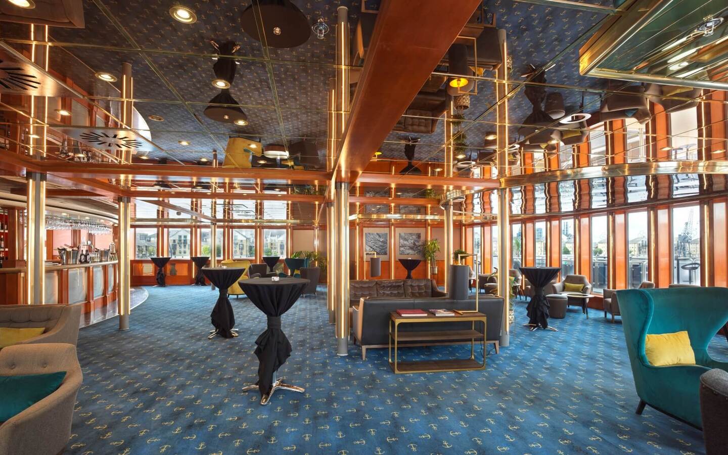 Waterfront hotel lounge with mirrored ceiling, brass and wood accents, anchor-pattern blue carpet, bar, cocktail tables and plush seating.