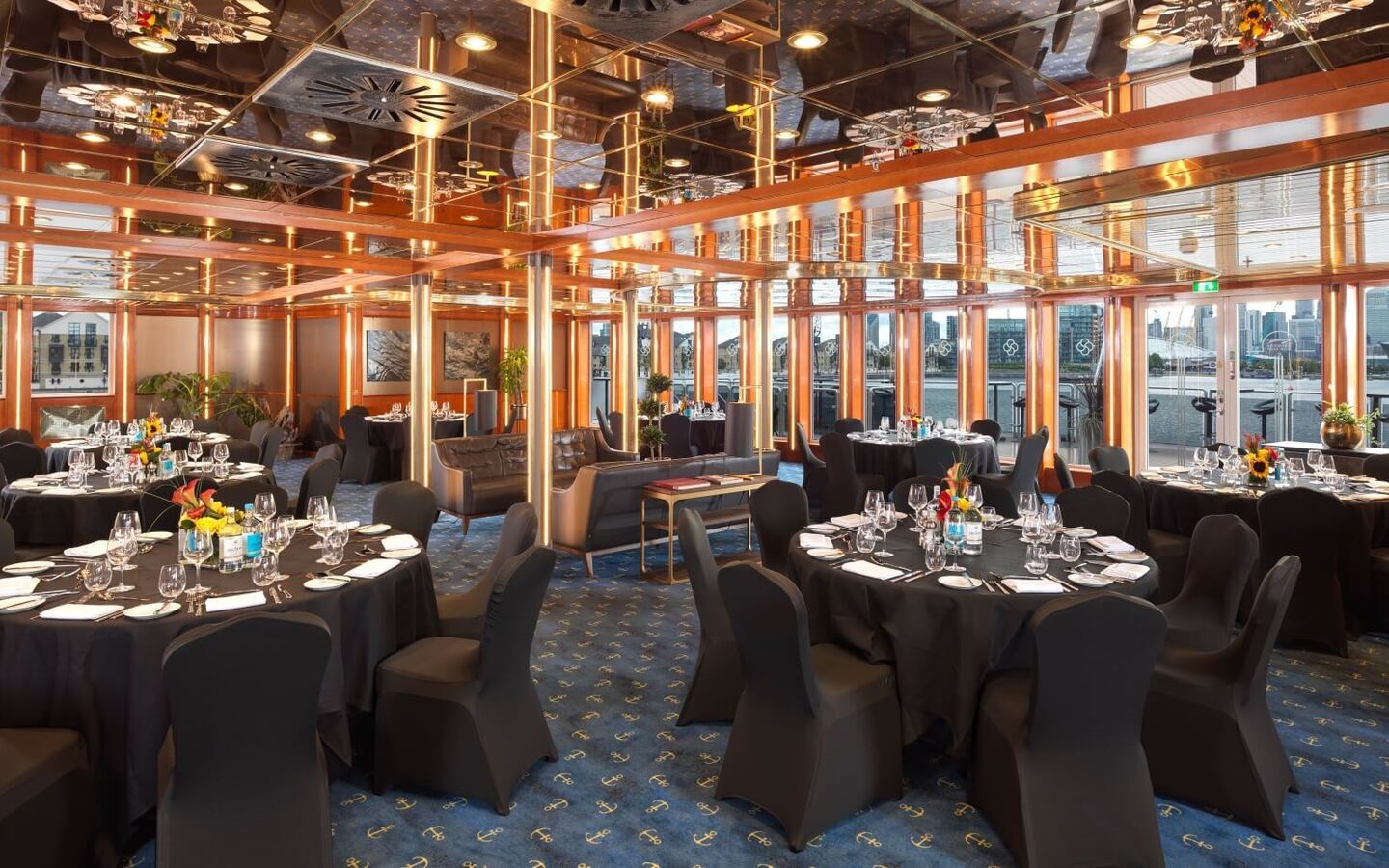 Elegant waterfront banquet room with round black-clothed tables, formal place settings, mirrored ceiling and city skyline views.
