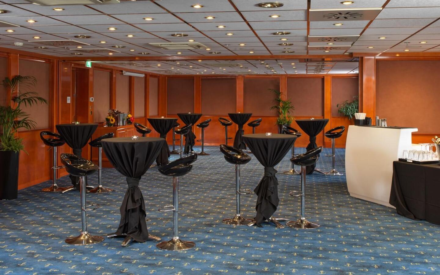 Wood-paneled hotel event room set for a cocktail reception with black-draped high tables, glossy bar stools, bar station and wine buckets.