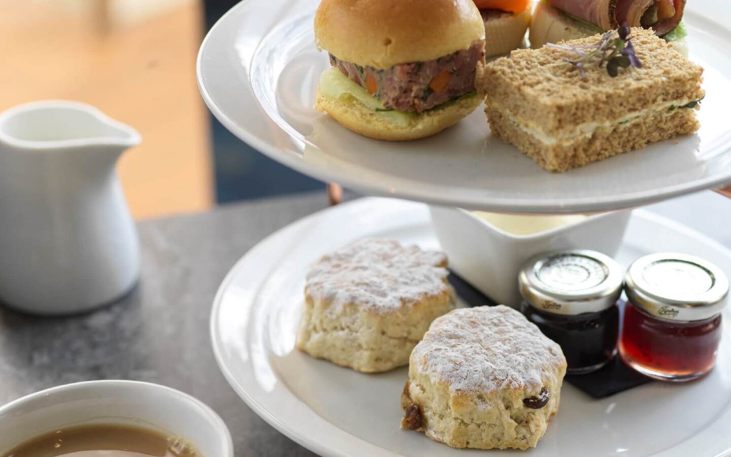 Tiered afternoon-tea stand with powdered scones, jam jars, tea and assorted sandwiches: brioche slider, smoked salmon, roast beef roll.