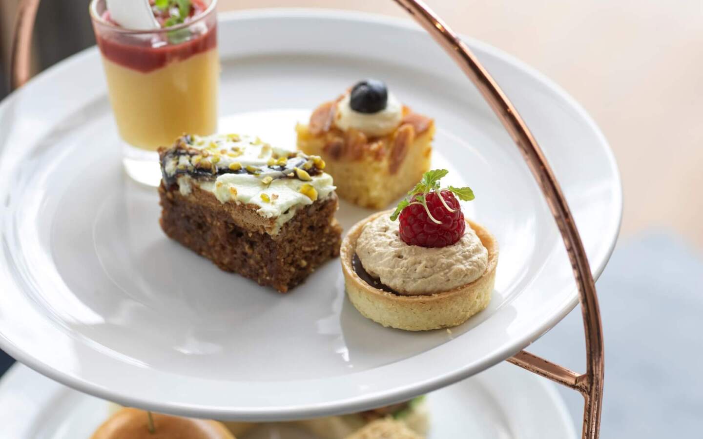 Afternoon tea pastries on a white tiered stand: raspberry tart, pistachio cake, almond square and custard shot