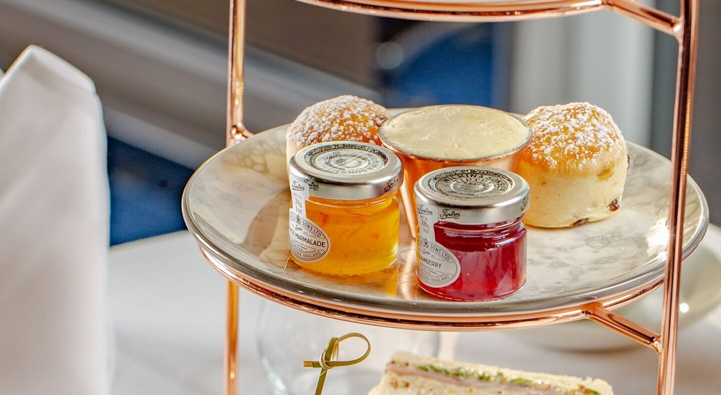 Three-tiered tea stand with freshly baked scones dusted with sugar, clotted cream and jars of marmalade and strawberry jam.
