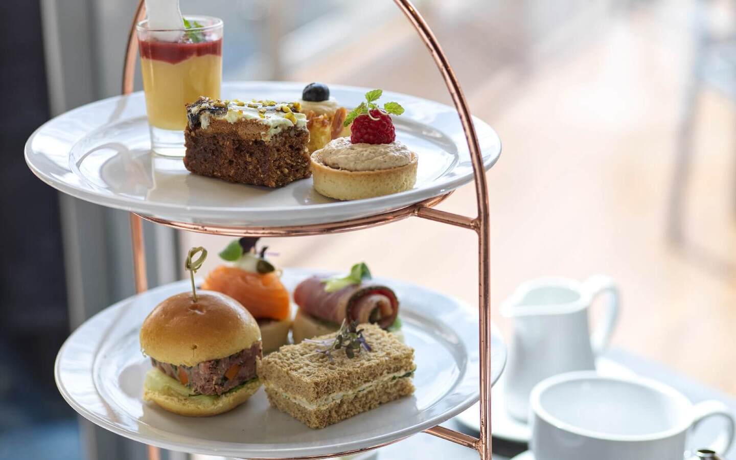 Three-tier afternoon tea stand with mini sandwiches and slider, smoked salmon rolls, petit tarts and desserts on copper frame