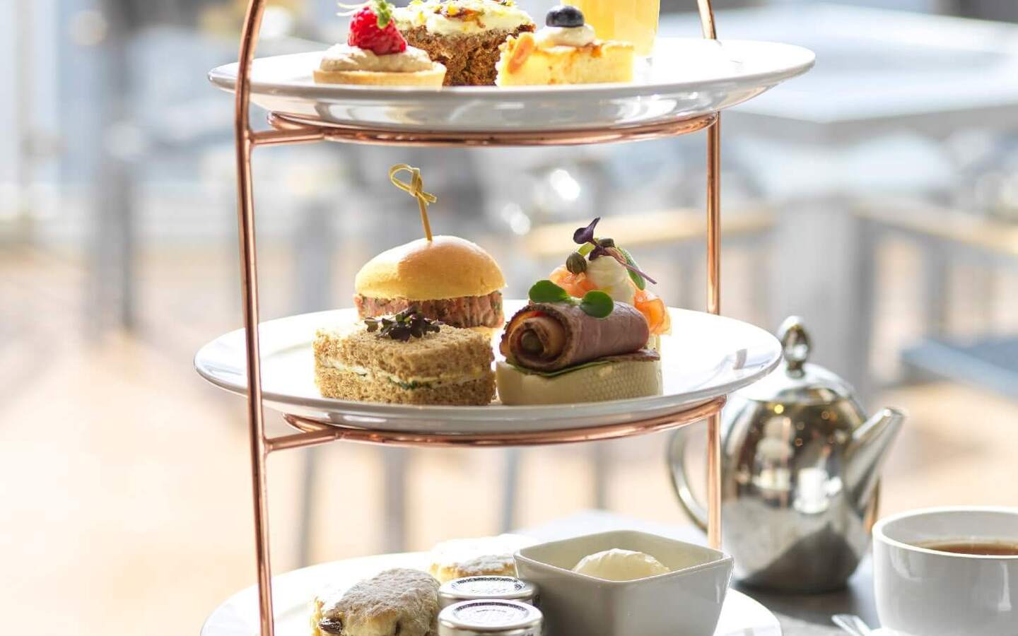Three-tiered afternoon tea stand with scones, finger sandwiches, pastries and dessert glass beside teapot on sunlit terrace.