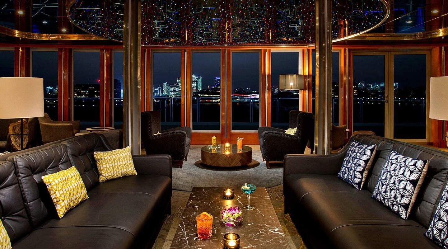 Hotel lounge at night with black leather sofas, patterned cushions, cocktails on a marble table and city skyline views.