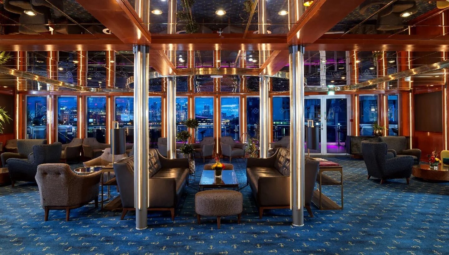 Upscale hotel lounge with leather sofas, armchairs, mirrored ceiling, metal-lit columns, anchor carpet and waterfront view.