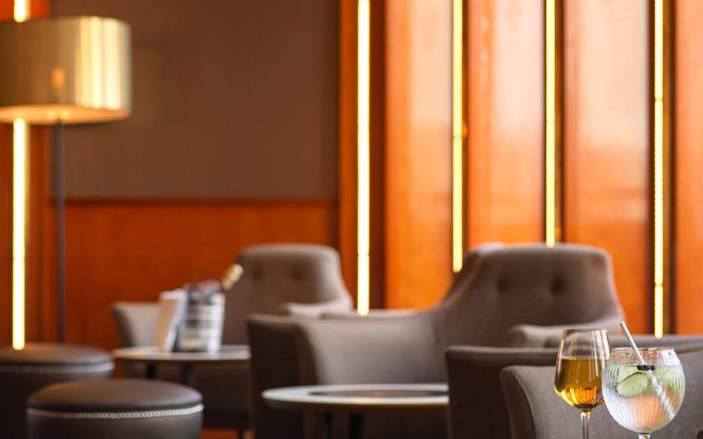 Warm wood-paneled hotel lounge with tufted armchairs, ottomans, round tables, vertical ambient lighting and two cocktails