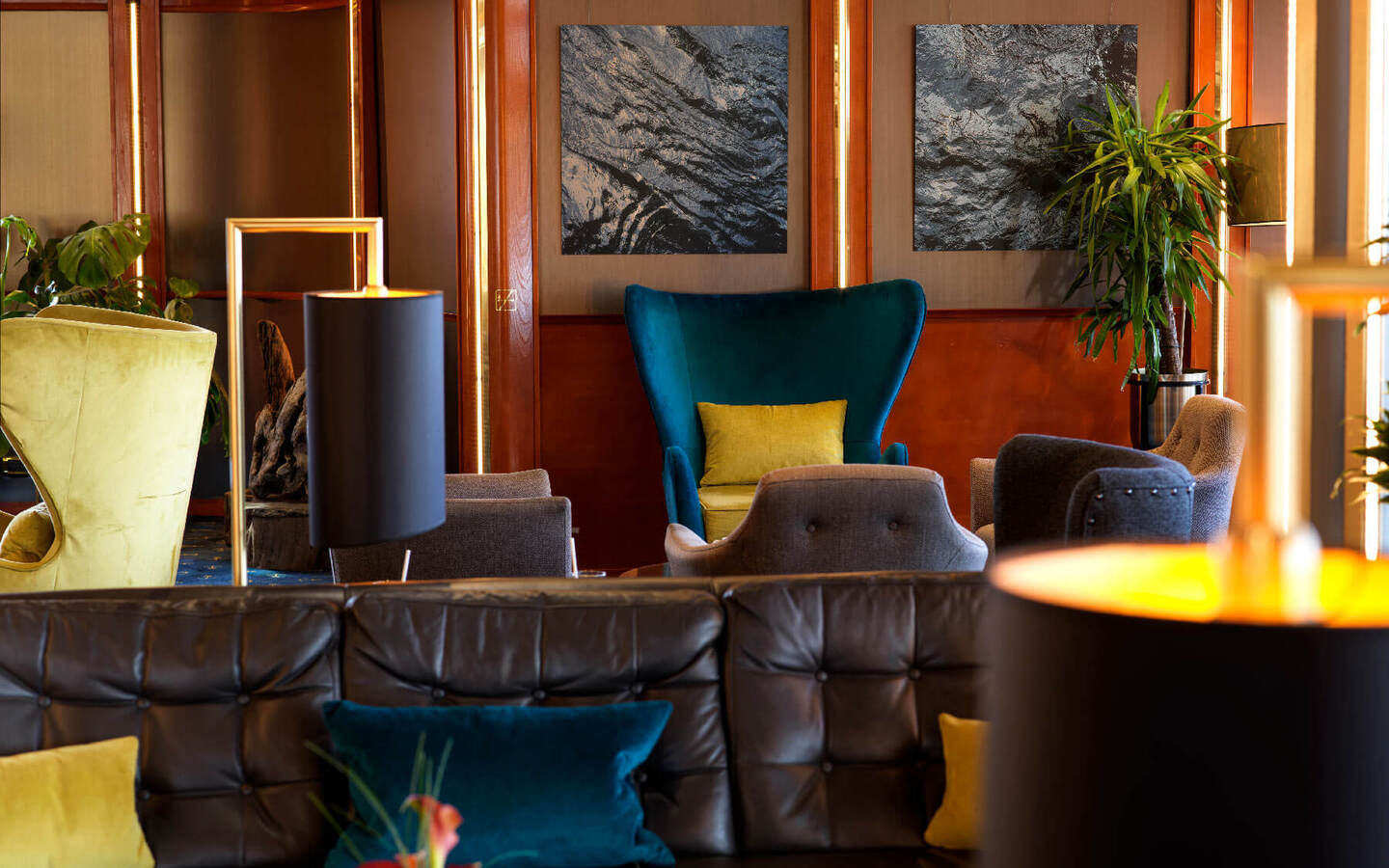Cozy hotel lounge with leather sofa, teal velvet wingback chair, colorful pillows, wood paneling, modern lamps and plants.