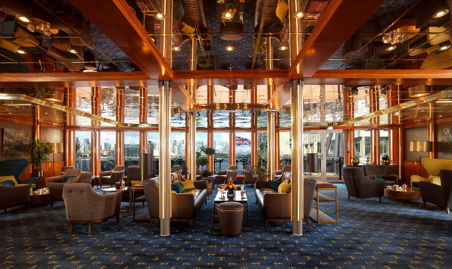 Warm hotel lounge with plush seating, mirrored ceiling, wood accents and nautical carpet, large windows with river views.